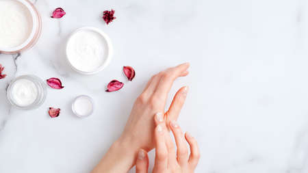 Hand skin care concept. Top view female hands applying organic moisturizing hand cream, jars with cosmetic cream and pink petal on marble table. Natural organic beauty productsの写真素材