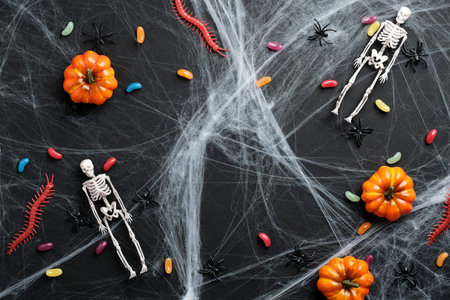 Black halloween background with spider web, colorful candies, pumpkins, skeletons. Flat lay, top...