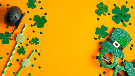 Happy St. Patrick's Day frame of shamrock leaf clovers, pot of gold, party glasses, drinking straws on orange background. Saint Patricks Day flat lay compositionの写真素材