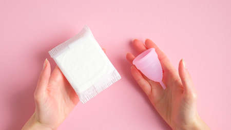 Female hands holding sanitary pad and menstrual cup over pink background. Different types of menstrual hygiene products comparison. Critical days, menstruation cycle, female healthcare conceptの写真素材
