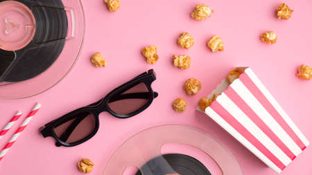 Watching movie with popcorn concept. Flat lay composition with 3d glasses, pop corn in red and white cardboard box, reels of film and drink straws on pink background.の写真素材
