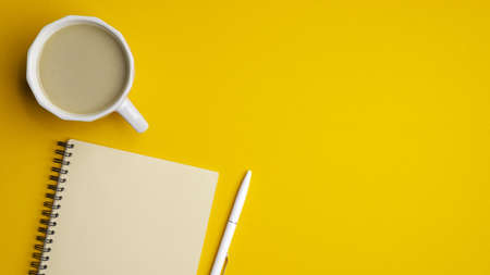 Modern office desk table with cup of coffee, notepad and pen on yellow background. Minimal style simple workspace. Top view with copy space, flat lay. Concept of business or educationの写真素材