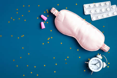 Flat lay composition with pink sleep eye mask, alarm clock, ear plugs, sleeping pills for insomnia on dark blue background with confetti. Sleep hygiene, insomnia treatment conceptの写真素材
