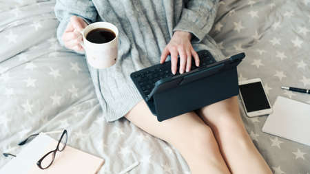 Young woman working on tablet with keyboard and drinking coffee in bed at home. Cozy, hygge style. Working home, stay safe, distance education conceptの写真素材