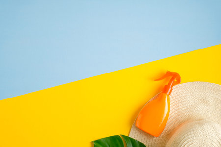 Sunblock spray bottle, beach hat and tropical leaf on blue and yellow background. Top view with copy...