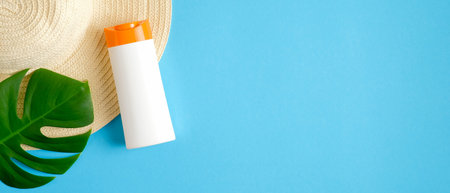 Blank sunscreen lotion bottle on blue background with tropical leaf and beach hat. Top view with...