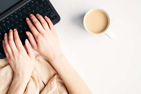Female hands typing a text on laptop keyboard top view. Flat lay modern minimal feminine workspace with blanket, coffee cup and notebookの写真素材