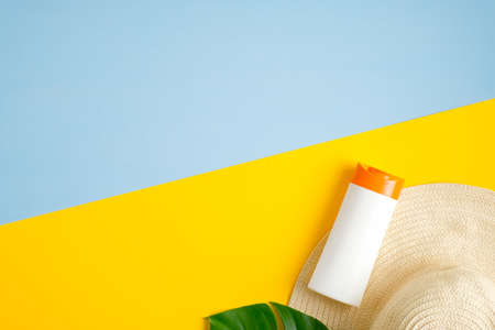 Sunscreen lotion bottles, green tropical leaf, female beach hat isolated on blue and yellow background. Sun protection, suntan cosmetics, skin care concept. Flat lay, top viewの写真素材