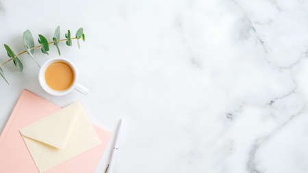 Home office desk table with pink notepad, pen, envelope, cup of coffee, eucalyptus leaf on marble background. Flat lay, top view. Elegant feminine workplace with stylish accessoriesの写真素材