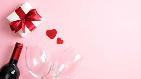 Valentines day greeting cart template. Flat lay white gift box with red ribbon bow, bottle of wine champagne, two glasses and hearts on pink background. Happy Valentines day, romantic dinner conceptの写真素材