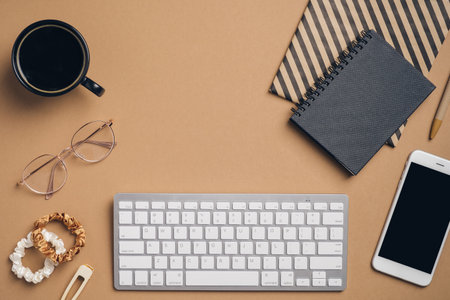 Top view home office desk table with computer keyboard, smartphone, paper notebooks, glasses, cup of coffee, female accessories on brown background. Feminine workspace concept.の写真素材