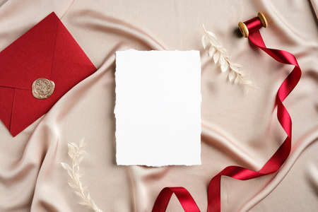 Elegant wedding stationery set top view. Flat lay blank invitation card mockup, red ribbon, envelope on beige satin.の写真素材