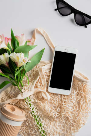 String bag with cup of coffee, flowers, smartphone mockup on white background. Elegant feminine accessories. Sustainable lifestyle concept.の写真素材