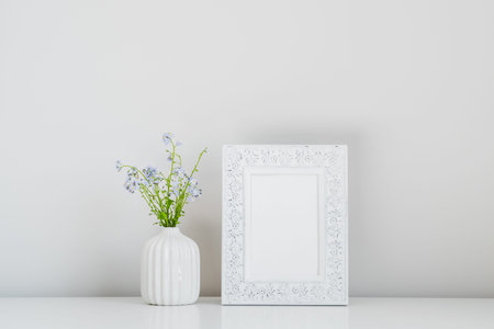 Photo frame mockup and vase with spring flowers on white table. Scandinavian home interior design.