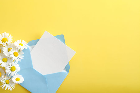Blue open envelope with blank paper card and chamomile flowers on yellow background. Flat lay composition, top view, copy space.の写真素材