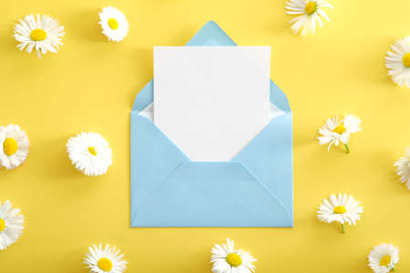 Blue envelope with white paper card mockup and chamomiles flowers on yellow background. Flat lay, top view, copy space. Love, romance concept.の写真素材