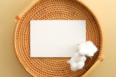 White invitation card mockup with cotton on rattan plate on beige backround. Flat lay, top view.