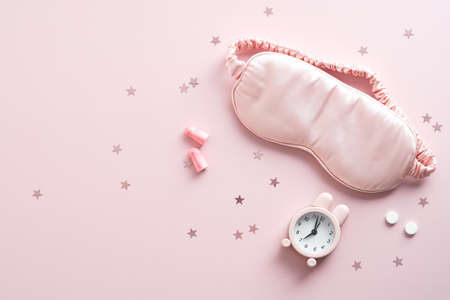 Flat lay sleep eye mask, alarm clock, earplugs, pills on pink background. Insomnia treatment accessoriesの写真素材