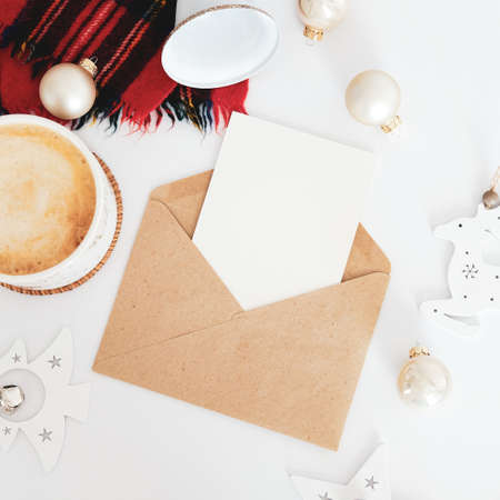 Craft paper envelope with Christmas letter mockup, cup of coffee, scarf, nordic Christmas decorations on white desk table. Scandinavian Christmas concept.の写真素材