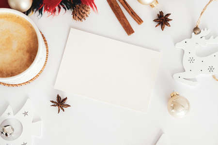Blank greeting card mockup, cup of hot chocolate, cinnamon sticks, scarf, vintage Christmas decorations on white desk table. Seasonal background, winter holidays composition. Flat lay, top viewの写真素材