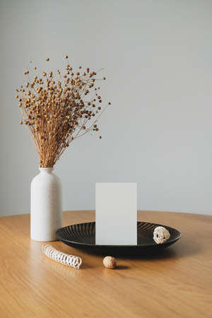 Vase with flax dry grass and blank paper card on wooden table. White wall on background. Still life nordic, Scandinavian home decorの写真素材