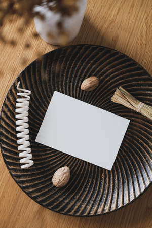 Blank paper card on plate with walnuts and autumn holiday decorations, vase of dried flowers on wooden table. Hygge, cozy home interior decorの写真素材