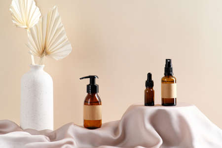 Set of natural cosmetics in amber glass packages on satin fabric round podium and vase of dried flowers on beige background.の写真素材