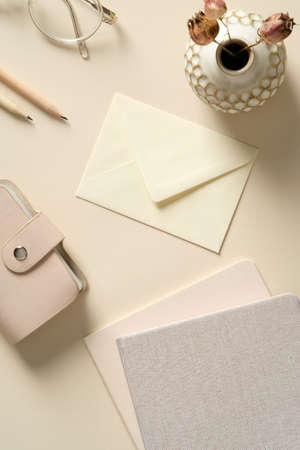 Stylish home office desk table with envelope, feminine accessories, paper notebooks, pencils on beige background. Flat lay, top view.の写真素材