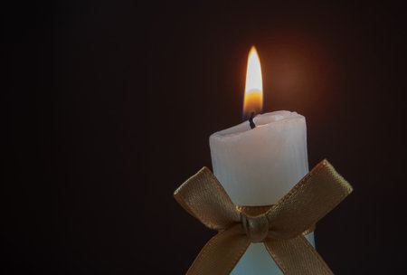 Burning ribbed pillar candle with golden satin silk ribbon tied bow on the dark background. Holiday home decor. Candlelight.の写真素材