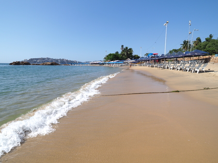 Sunbeds and umbrellas on sandy beach at bay of Acapulco city landscapes in Mexico and waves of Pacific Ocean with clear blue sky in 2018 hot sunny winter day, North America on March.のeditorial素材