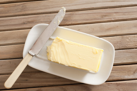 Pat of fresh farm butter on a butter dish with a knife to use as a spread or cooing ingredient, overhead view on a slatted wooden tableの素材 [FY31047200472]