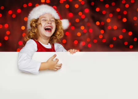 A portrait of a laughing sweet little curly girl in Santa hat looking out from behind empty blank, isolated against Christmas glitter close up. Holidays concept. Invitation. Funny kidsの写真素材