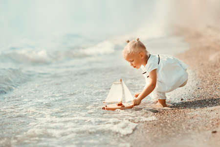 A cute little girl in white clothes playing with a toy ship on the beach on a warm sunny summer day. Holidays at sea. Funny kidsの写真素材