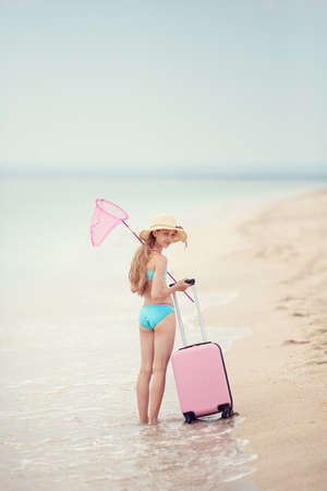 A teenage girl in a swimsuit and a straw hat carries a fish net and carries a pink suitcase along the sea beach. Rest. Holidays.の写真素材
