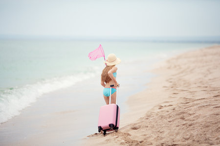 A teenage girl in a swimsuit and a straw hat carries a fish net and carries a pink suitcase along the sea beach. Rest. Holidays.の写真素材