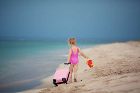 A little girl in a pink swimsuit is walking along the beach with a toy bucket and a pink suitcase. Holidays and recreationの写真素材