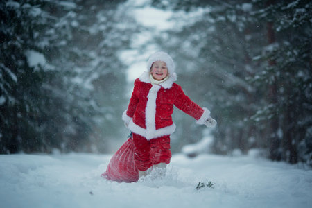Little funny child dressed in Santa Claus red costume bringing presents in winter snowy forest. Christmas Eveの写真素材