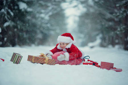 Little funny child dressed in Santa Claus red costume bringing presents in winter snowy forest. Christmas Eveの写真素材