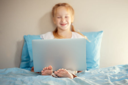 A cute girl sitting on the bed is watching something on a laptop. The concept of education