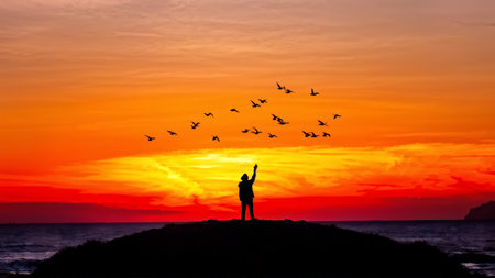 Silhouette of person with arms raised against sunset sky with birds in flight over water; dark sea below at dusk.の素材
