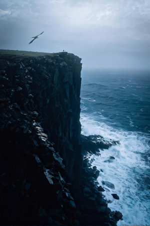 A bird in flight above a cliff with turbulent ocean waves below under a cloudy sky.の素材