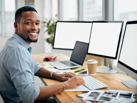 A cheerful African-American man working at a desk with multiple monitors, smiling at the camera in a professional office setting.の素材
