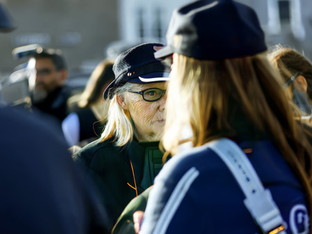 Two individuals engaged in a conversation outdoors during daylight. One is wearing glasses and a dark cap while the other has long blonde hair.の素材