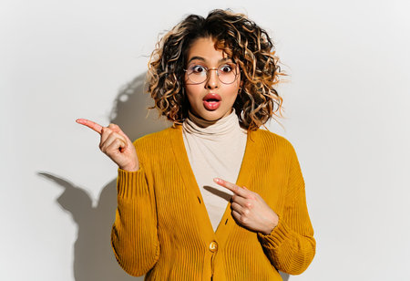 young african american woman with curly hair wearing glasses and amazed surprised looking up pointing fingers raised arms.young arms.の素材