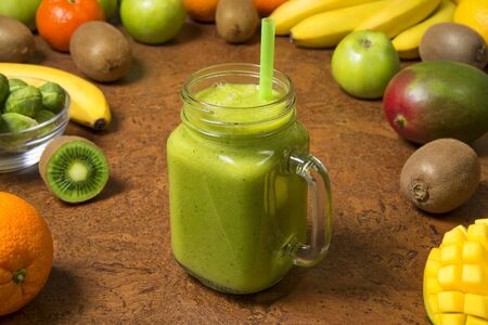 Green smoothie banana kiwi mango orange and apple in glass jar with fresh ingredients on a brown table. Selected focus.の写真素材