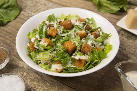 Caesar salad in a white bowl on an old rustic wooden table with leaves and a sauce boat and parmesan. Selected focus.の写真素材