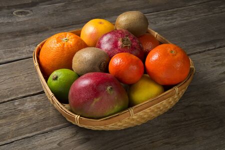 Fresh fruits of mango, kiwi, oranges, tangerines, pomegranates, lemon, lime and grapefruit in a basket on a rural wooden table.の写真素材