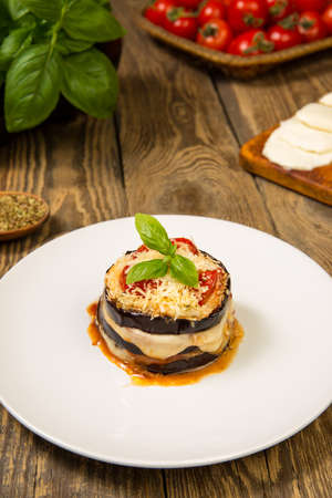 Baked eggplant and tomatoes mozzarella and parmesan stacks with ingredients on a rustic wooden table. Close up. Selected focus.の写真素材