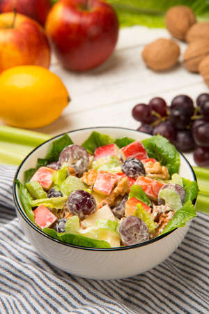 Waldorf salad of apples, celery stalks, walnuts, grapes, lettuce in a white salad bowl with ingredients on an old rustic white wooden table. selected focus.の写真素材