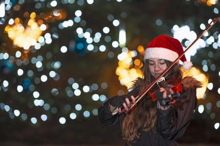 girl playing the violin in front of a Christmas tree. The lights on the Christmas tree are blurred and form a pleasant background. The girl is wearing a Christmas hat.の写真素材